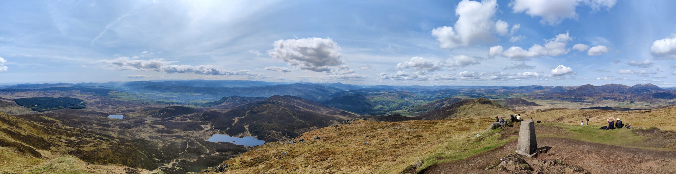 A wide angle panorama looking around the summit under a blue sky. In the distance, a mountain horizon, forestry plantations, lochans, valleys, and happy hikers enjoying their lunch