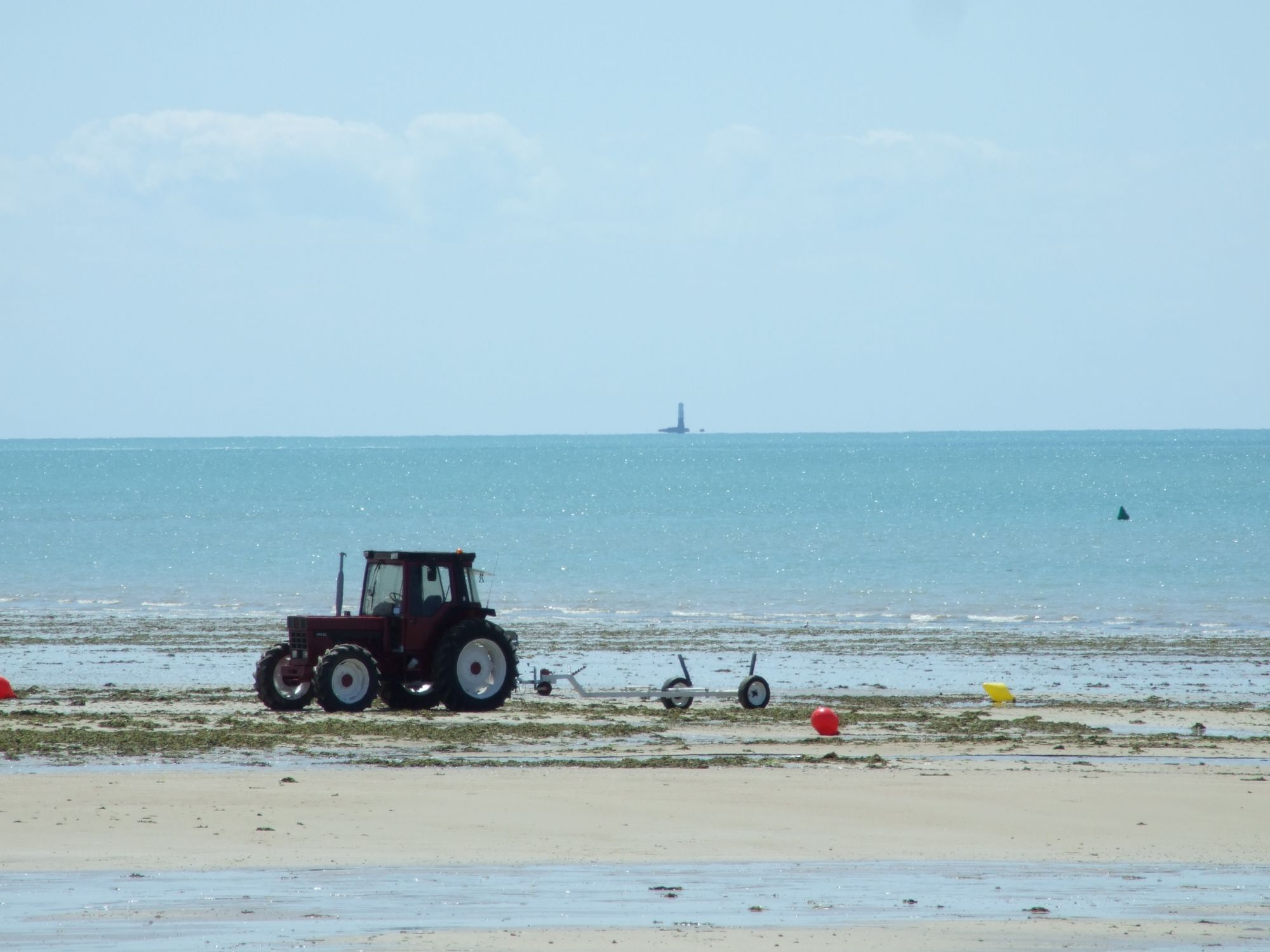 A tractor parked on a beach, looking out to sea with a lighthouse in the distance, almost floating like a mirage.