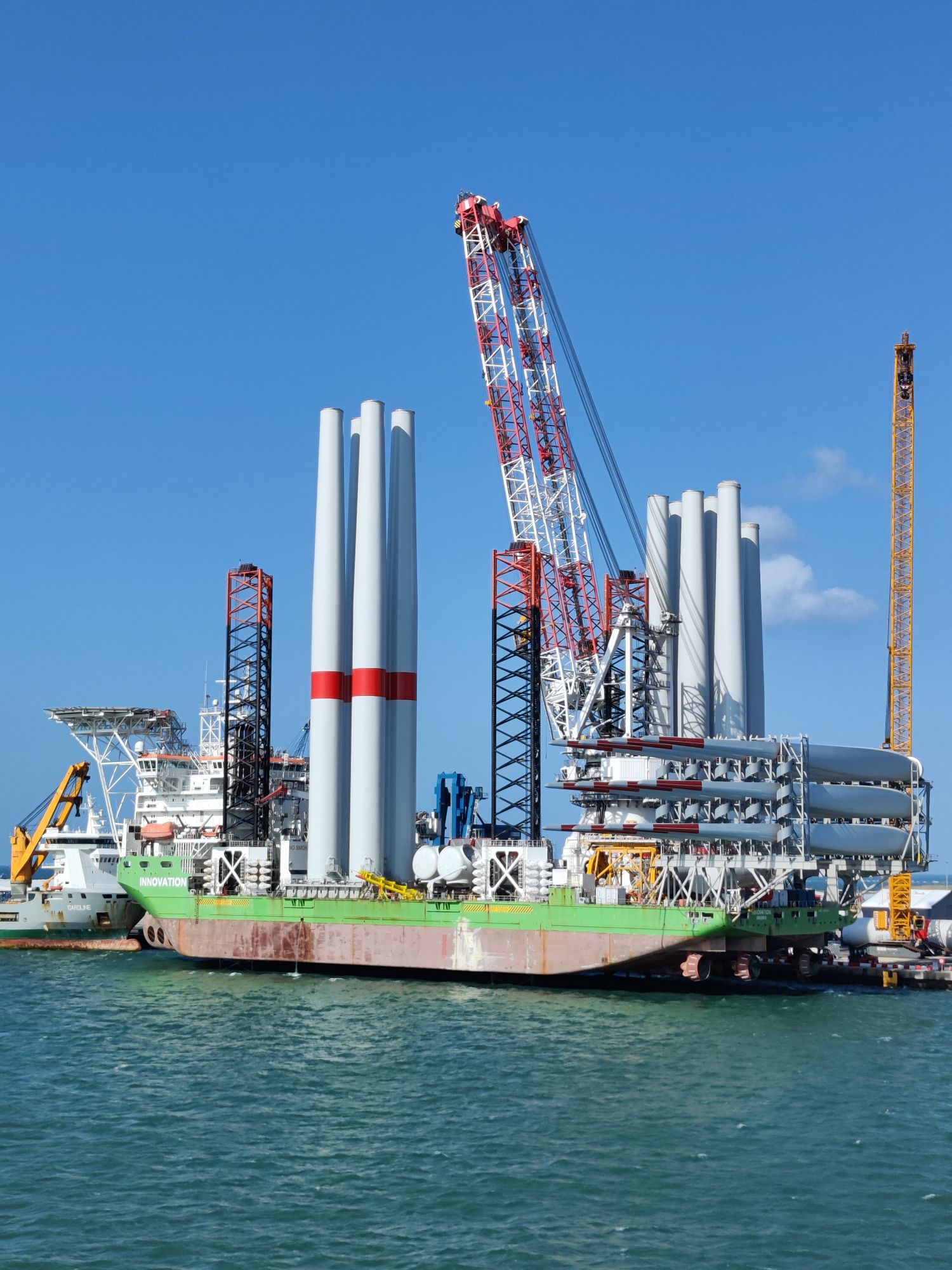 Some large wind turbines being loaded onto a barge that has 3 pylons used as stilts.